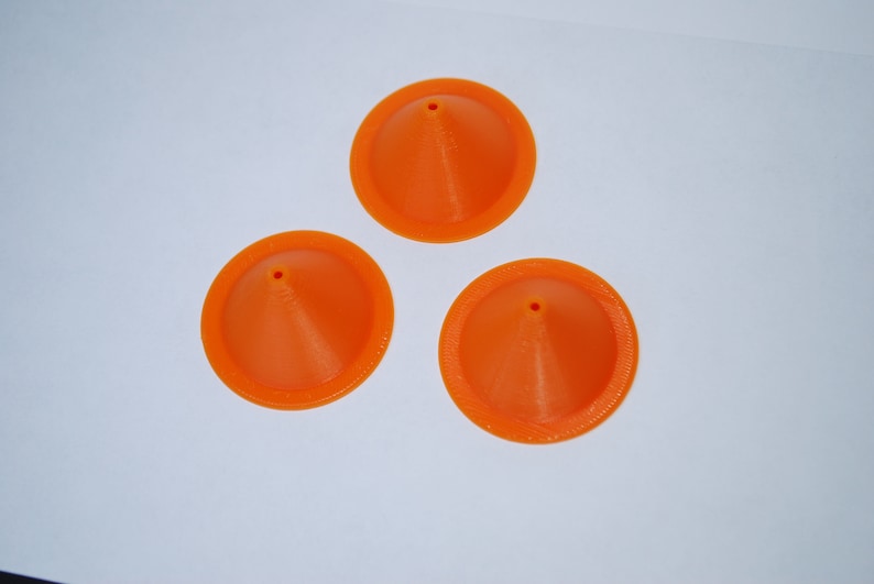 Pu&ograve; includere: Tre imbuti di plastica arancione con un piccolo foro sulla punta. Gli imbuti sono su una superficie bianca.