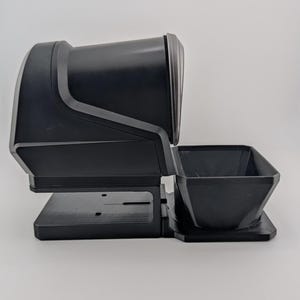 Op de afbeelding: Een zwart 3D-geprint kunststof accessoire met een cilindrische behuizing op een rechthoekige basis en een kleinere, open vierkante bak aan de voorzijde, alles verbonden door een plat, solide zwart platform als één geheel.
