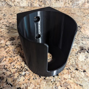 May include: A black, 3D-printed wall mount with a curved design. It features two small holes near the top and a larger circular opening at the bottom. The mount is designed to hold an item, such as a phone or a small speaker.