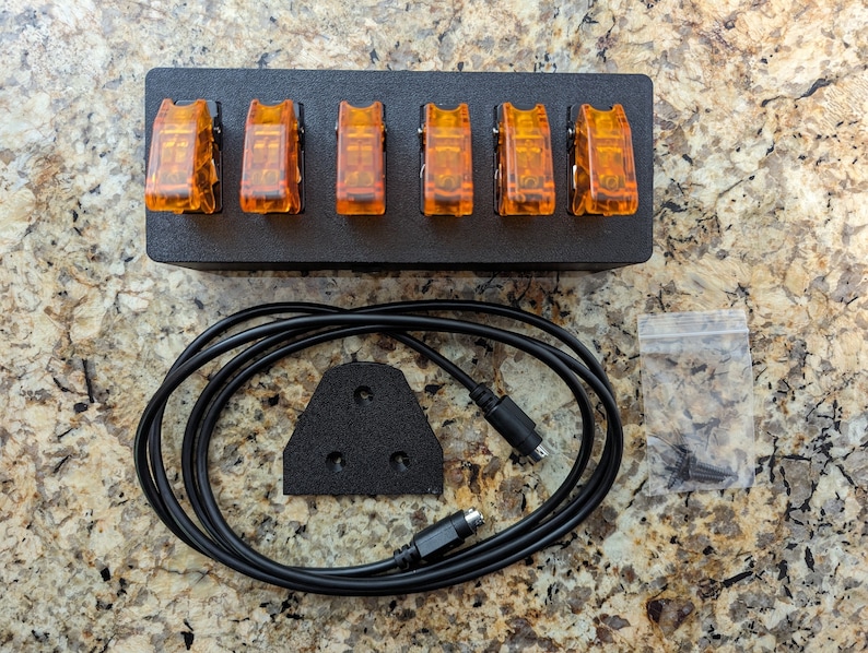 Puede incluir: Una caja negra con seis interruptores de palanca naranja, un cable negro con un conector en cada extremo, una placa de montaje negra con tres agujeros y una peque&ntilde;a bolsa de pl&aacute;stico que contiene tornillos.