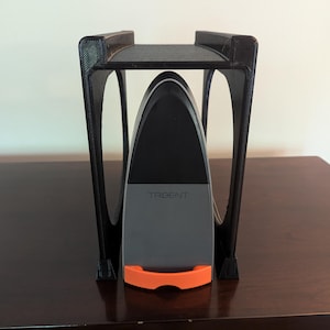 May include: A black plastic stand with a curved top holds a gray and black device with the word "TRIDENT" printed on it. The device has an orange handle.