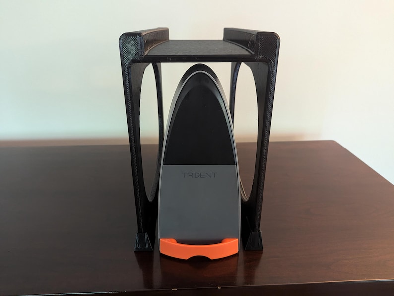 May include: A black plastic stand with a curved top holds a gray and black device with the word "TRIDENT" printed on it. The device has an orange handle.