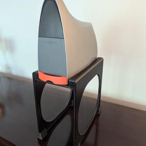 May include: A gray and silver 3D printer with the word "TRIDENT" printed on the front, sitting on a black plastic stand with four legs.