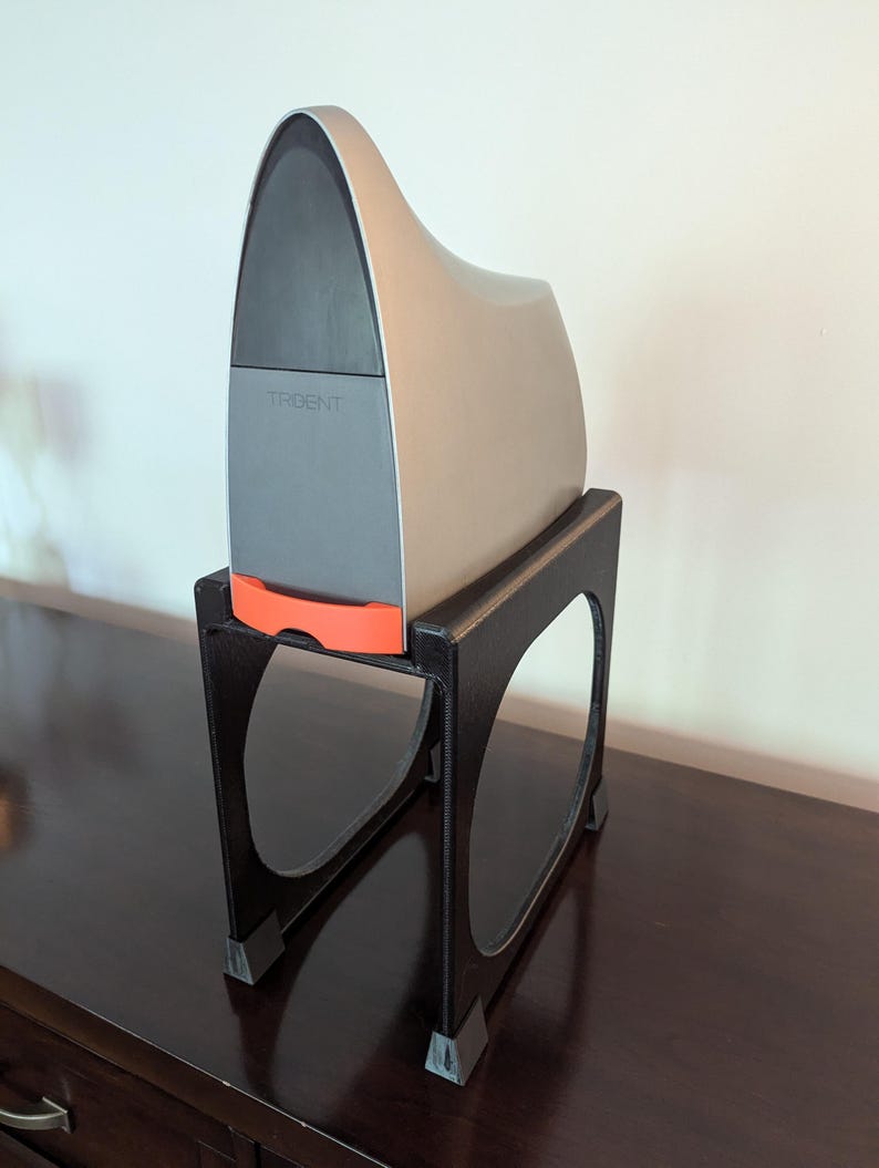 May include: A gray and silver 3D printer with the word "TRIDENT" printed on the front, sitting on a black plastic stand with four legs.