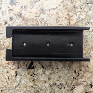 Puede incluir: Soporte de teléfono de plástico negro con tres agujeros para tornillos. El soporte está diseñado para ser fijado a una pared u otra superficie.