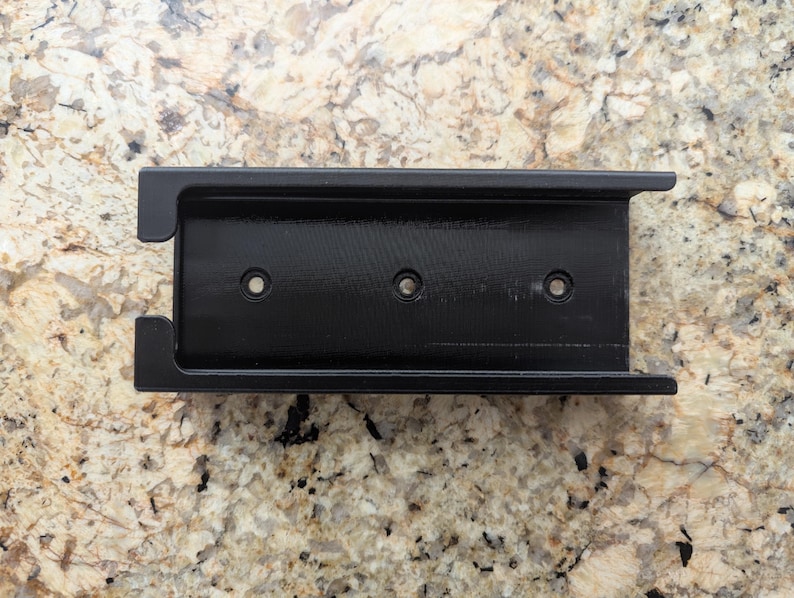 May include: Black plastic phone mount with three holes for screws. The mount is designed to be attached to a wall or other surface.