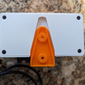 May include: A white rectangular device with a bright orange plastic stand on the back. The device has four black screws on the sides and a black cable attached to the bottom.