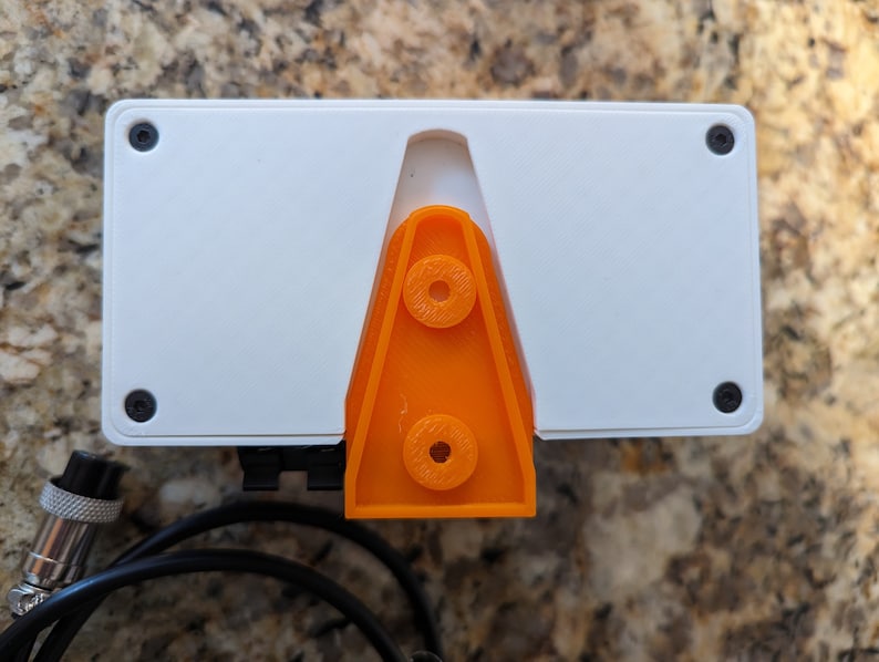 May include: A white rectangular device with a bright orange plastic stand on the back. The device has four black screws on the sides and a black cable attached to the bottom.