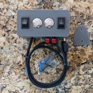 May include: A gray box with two silver buttons and two black toggle switches. The box has red and black speaker wire connectors and a USB cable. The box is on a granite countertop.