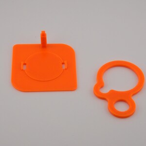 Pu&ograve; includere: Due componenti in plastica arancione brillante. Uno &egrave; un quadrato con un inserto circolare e una linguetta rialzata. L'altro &egrave; un anello con un anello pi&ugrave; piccolo attaccato. Entrambi sono su sfondo bianco.