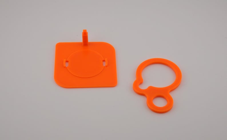 Pu&ograve; includere: Due componenti in plastica arancione brillante. Uno &egrave; un quadrato con un inserto circolare e una linguetta rialzata. L'altro &egrave; un anello con un anello pi&ugrave; piccolo attaccato. Entrambi sono su sfondo bianco.