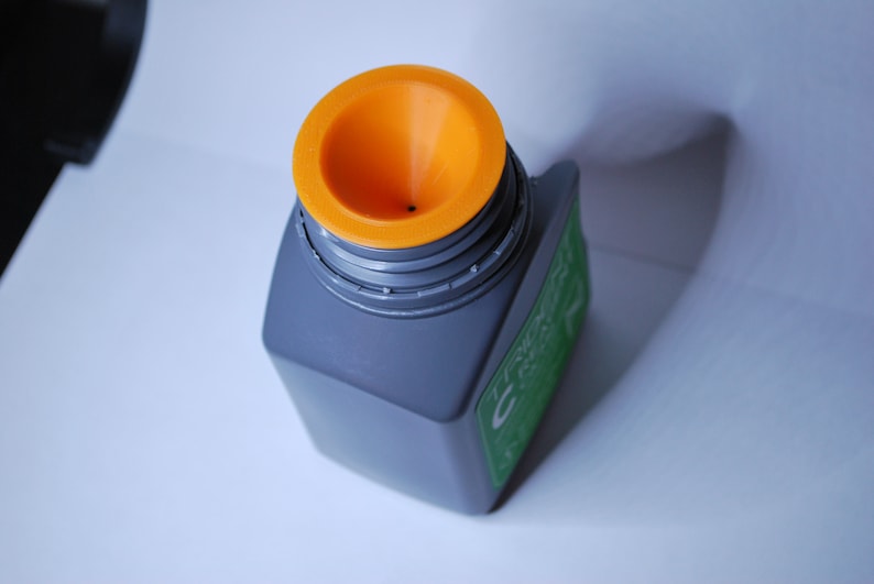 Pu&ograve; includere: Una bottiglia di plastica grigia con un'etichetta verde e un tappo a forma di imbuto arancione. La bottiglia ha un piccolo foro al centro del tappo.