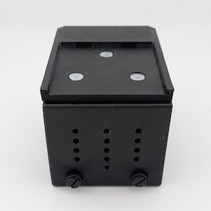 May include: A black rectangular box with a flat top and three white circular accents. The front of the box has three vertical rows of holes and two knobs at the bottom. The box is likely a component or housing for electronics.