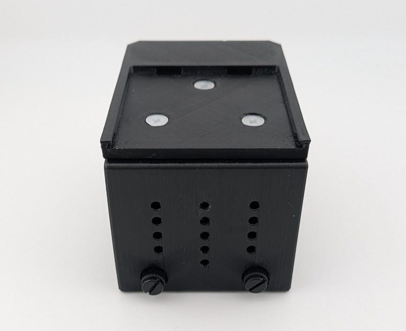 May include: A black rectangular box with a flat top and three white circular accents. The front of the box has three vertical rows of holes and two knobs at the bottom. The box is likely a component or housing for electronics.