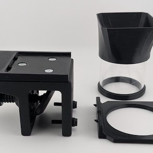 May include: A set of black 3D-printed components, including a mounting bracket with a screw, a clear cylindrical container with a black top, and a circular frame. The items are arranged on a white surface.