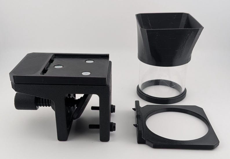 May include: A set of black 3D-printed components, including a mounting bracket with a screw, a clear cylindrical container with a black top, and a circular frame. The items are arranged on a white surface.