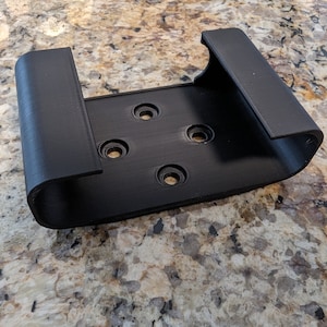 May include: A black, rectangular bracket with rounded edges and four circular holes. The bracket has two curved arms extending upwards. The object is made of a matte black material.