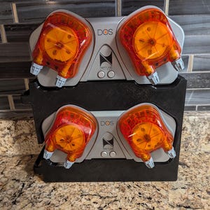 May include: Two silver and black DOS peristaltic pumps with orange heads are mounted on a black stand. The pumps are used for dosing liquids in aquariums.