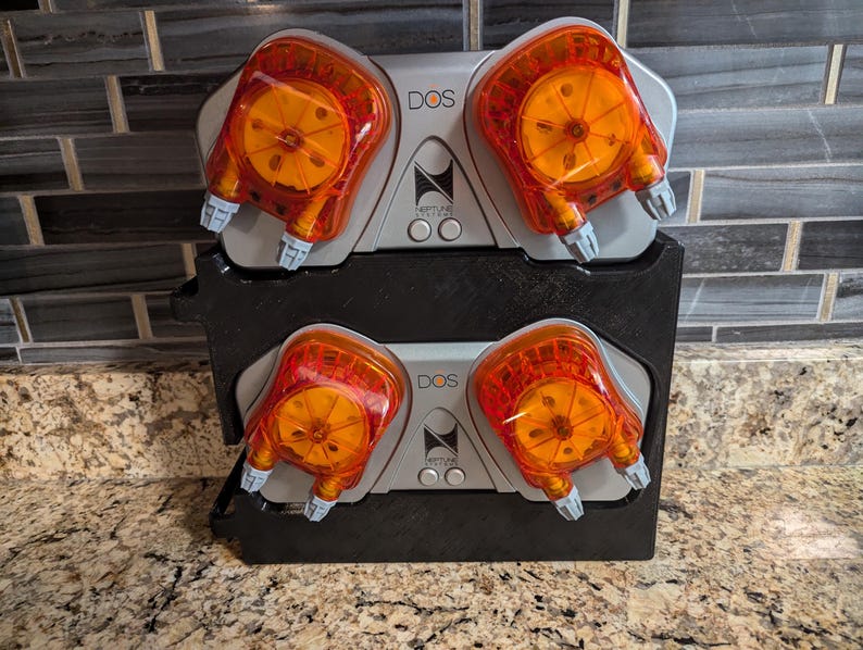May include: Two silver and black DOS peristaltic pumps with orange heads are mounted on a black stand. The pumps are used for dosing liquids in aquariums.
