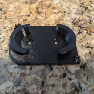 May include: A black plastic 3D printed wall mount with two circular cutouts for holding objects.