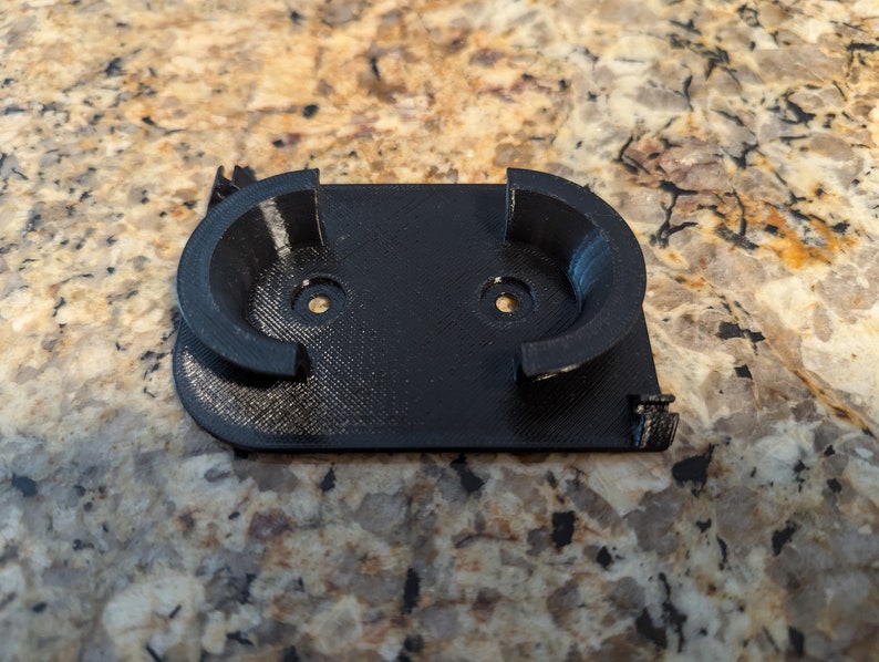 May include: A black plastic 3D printed wall mount with two circular cutouts for holding objects.