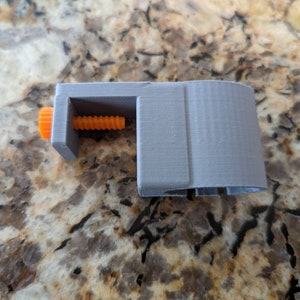 May include: A gray 3D printed clamp with an orange knob and a threaded screw. The clamp is designed to hold objects securely.