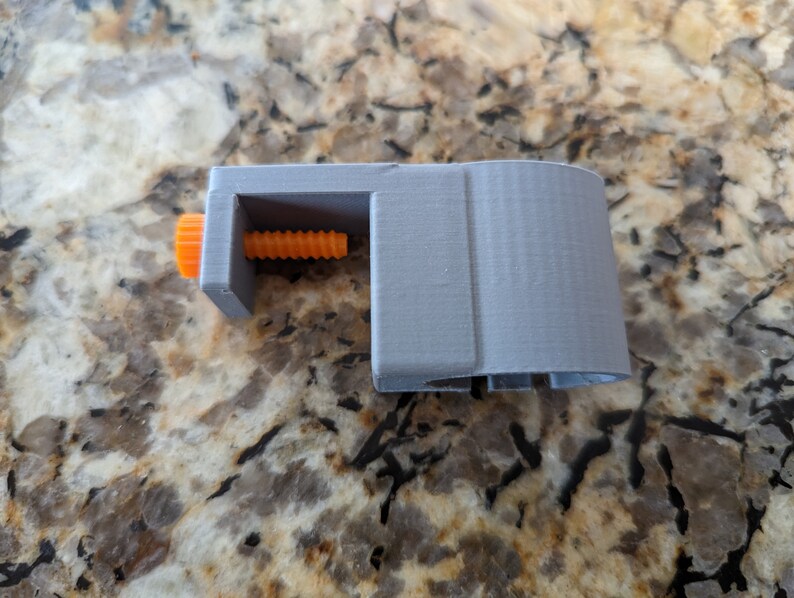 May include: A gray 3D printed clamp with an orange knob and a threaded screw. The clamp is designed to hold objects securely.