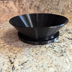 May include: A black plastic funnel with a wide opening and a narrow spout, sitting on a black plastic base. The funnel is designed for pouring liquids or powders into containers with small openings.