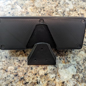 Puede incluir: Caja rectangular negra impresa en 3D con un soporte triangular. La caja tiene cuatro agujeros de tornillo visibles en la parte superior e inferior.