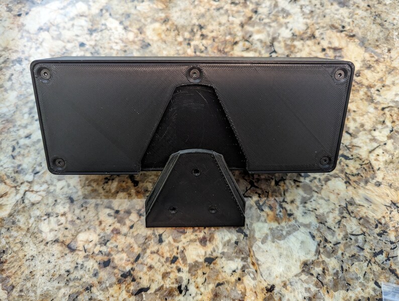 Puede incluir: Caja rectangular negra impresa en 3D con un soporte triangular. La caja tiene cuatro agujeros de tornillo visibles en la parte superior e inferior.
