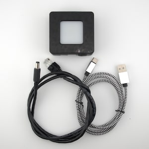 May include: A black square device with a central illuminated square, accompanied by a black power cable, an Ethernet cable, and a USB-C to USB-A cable with a black and white pattern. The items are arranged on a white surface.