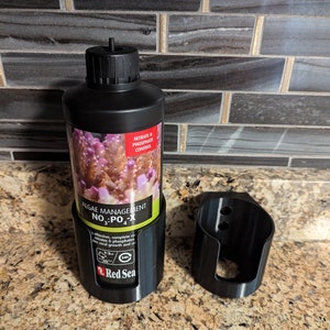 May include: A black plastic bottle with a red label that says "Red Sea" and "Algae Management NO3:PO4-X". The bottle is in a black plastic holder. The bottle has a red and white label with the text "Nitrate & Phosphate Control".