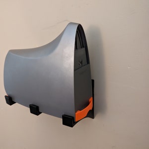 Könnte beinhalten: Ein grauer wandmontierter Druckerhalter mit einem orangefarbenen Entriegelungshebel. Der Halter hat eine schwarze Basis und drei schwarze Montagehalterungen.