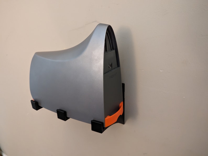 May include: A gray wall-mounted printer holder with an orange release lever. The holder has a black base and three black mounting brackets.