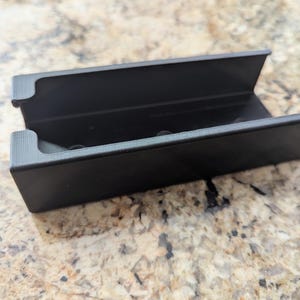 May include: A black plastic storage case with a rectangular shape and a slot on the top. The case has a smooth surface and is designed to hold small items.