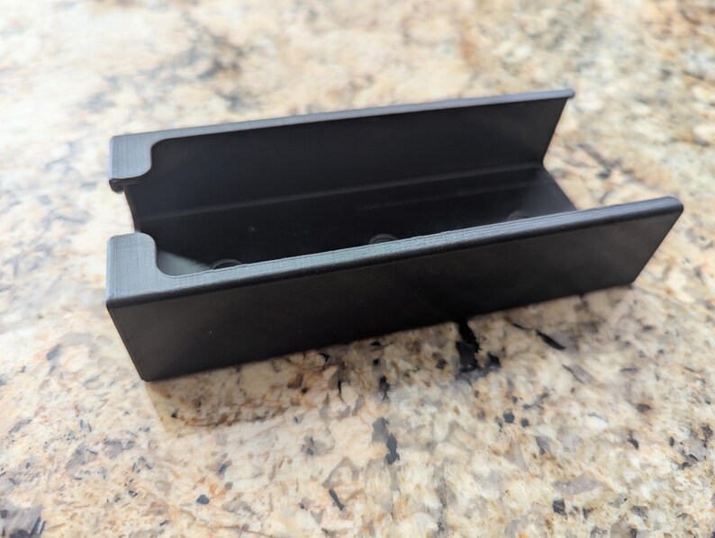 May include: A black plastic storage case with a rectangular shape and a slot on the top. The case has a smooth surface and is designed to hold small items.