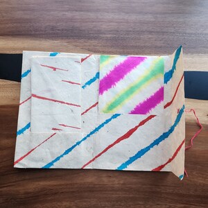 May include: A handmade paper folder with two pockets and a decorative tie-dye paper insert. The folder has a striped pattern in red and blue.
