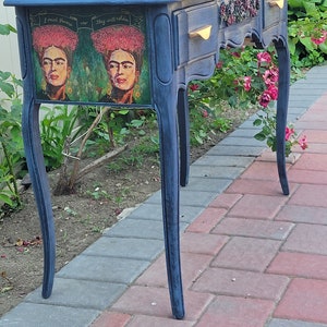 May include: A blue painted vanity table with two drawers and a distressed finish. The table features a decorative floral design on the drawers and a printed image of Frida Kahlo with the text "I paint flowers so they will not die".
