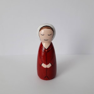 May include: Hand-painted wooden doll wearing a red dress and a white bonnet. The doll has brown hair and closed eyes.