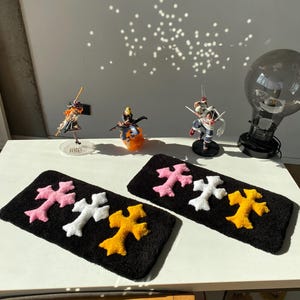 May include: Two rectangular black tufted rugs, each featuring three cross-shaped appliques in pink, white, and yellow. The rugs have a textured, plush surface and are arranged side-by-side on a white surface, showcasing a consistent design.
