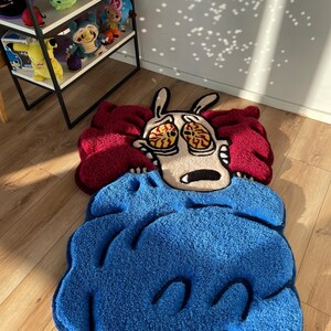 May include: A blue and red rug with a cartoon character of a dog with big eyes and a worried expression. The dog is under a blue blanket and has a red and white head.