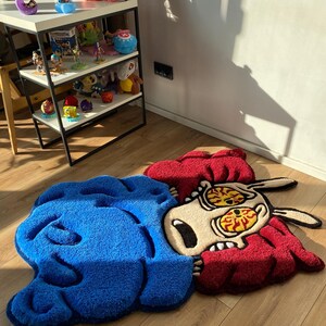 May include: A blue and red cartoon rug with a distressed look. The rug features a character with large eyes and a distressed expression. The rug is made of a soft, plush material.