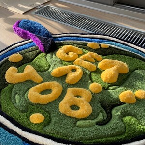 May include: A round, green and blue rug with the word 'GET YOUR' written in yellow, fluffy letters. The rug has a white and black border.