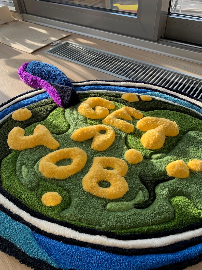 May include: A round, green and blue rug with the word 'GET YOUR' written in yellow, fluffy letters. The rug has a white and black border.