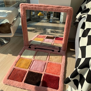 May include: A pink makeup palette-shaped rug with a large mirror. The rug features a grid of colorful squares in shades of beige, yellow, pink, orange, and red. The mirror and rug are made of a soft, plush material.