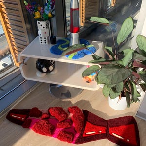 May include: A red and pink fuzzy rug with a geometric design. The rug is on a wooden floor in front of a white shelf with a lava lamp, a vase of flowers, and a black and white polka dot ceramic object.