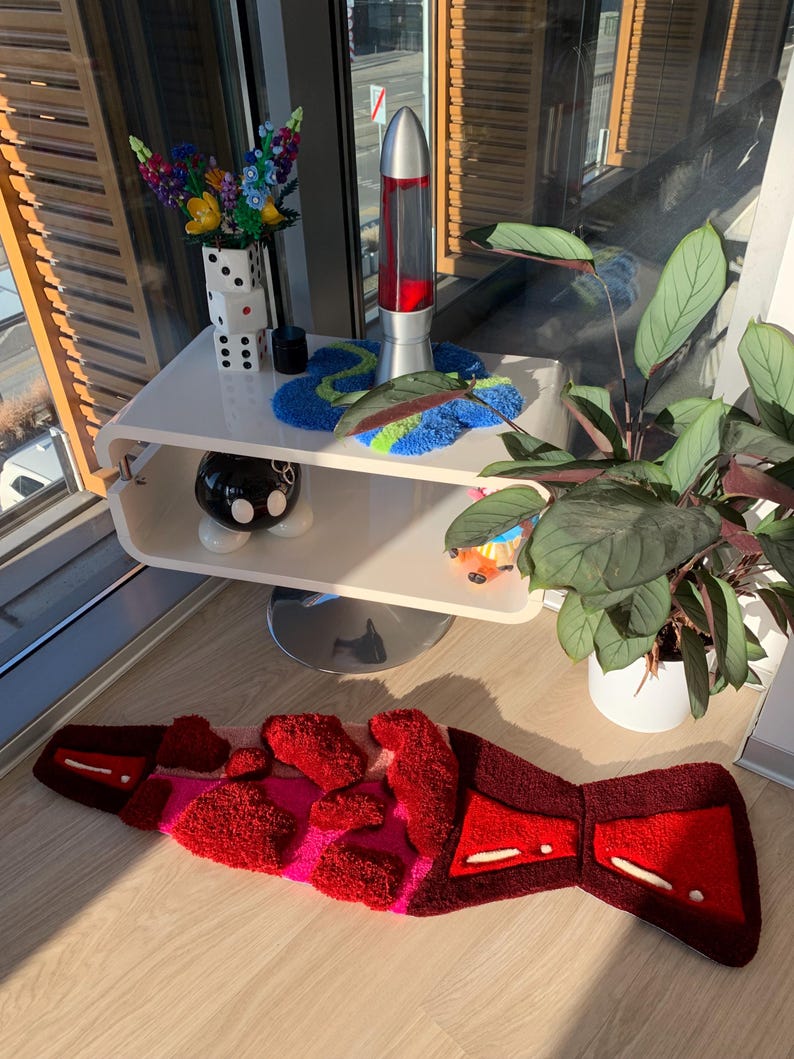 May include: A red and pink fuzzy rug with a geometric design. The rug is on a wooden floor in front of a white shelf with a lava lamp, a vase of flowers, and a black and white polka dot ceramic object.