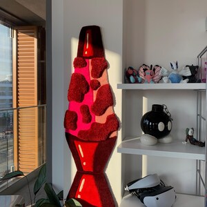 May include: A large, red and pink lava lamp rug hanging on a white wall. The rug is made of fluffy, textured material and has a realistic lava lamp design.