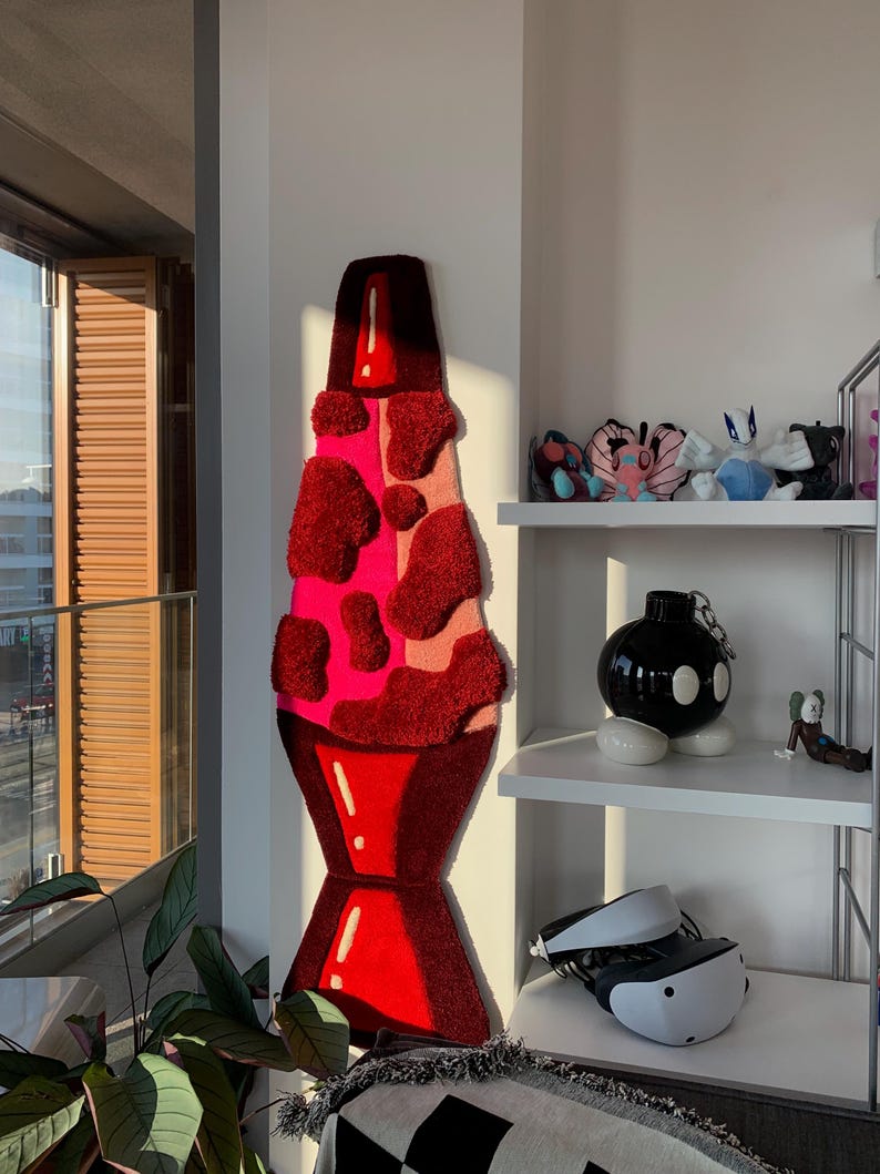 May include: A large, red and pink lava lamp rug hanging on a white wall. The rug is made of fluffy, textured material and has a realistic lava lamp design.
