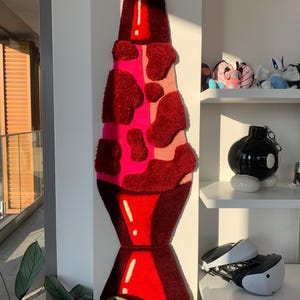 May include: A red and pink lava lamp rug with a white outline. The rug is made of fluffy, textured material and is shaped like a traditional lava lamp.